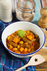 Butternut squash, pumpkin and bean stew with beef sausages. White bowl, dark blue tablecloth, wooden board.
