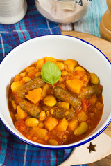 Butternut squash, pumpkin and bean stew with beef sausages. White bowl, dark blue tablecloth, wooden board.