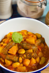 Butternut squash, pumpkin and bean stew with beef sausages. White bowl, dark blue tablecloth, wooden board.
