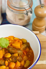Butternut squash, pumpkin and bean stew with beef sausages. White bowl, dark blue tablecloth, wooden board.