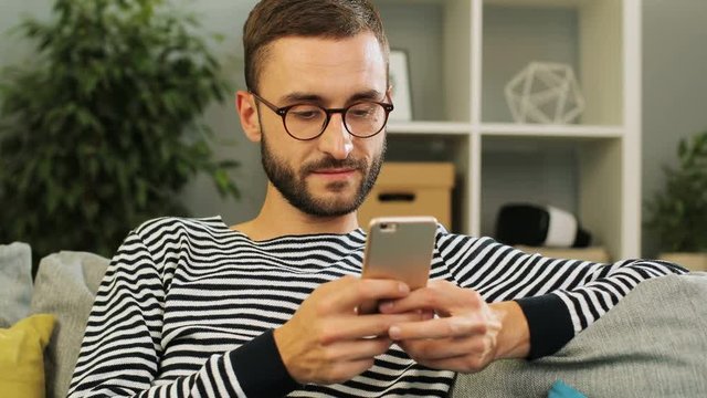 Young caucasian man in glasses sitting on the sofa using mobile phone to olay games, at home on living background. Indoor shot. Close up.