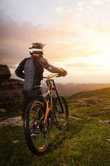 The rider in full protective equipment on the mtb bike is riding toward the sunset in the rays of...