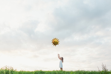 Little girl reaching out to big sun balloon against cloudy sky