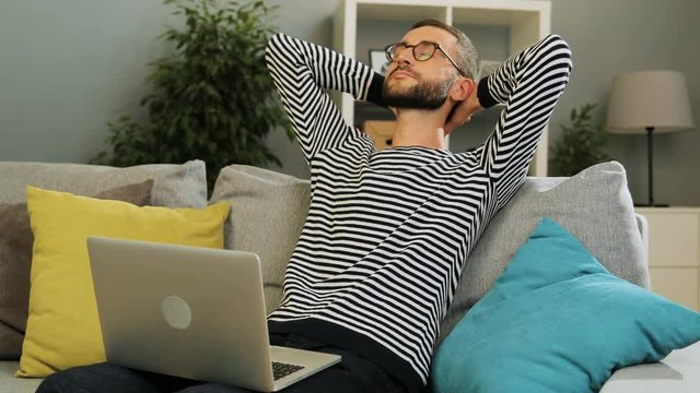 Handsome caucasian man in glasses sitting on the sofa typing on the laptop and then puttinh hends over his head to relax after hard work at the home background. Indoor shot.