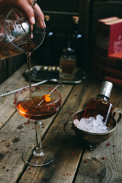 male barkeeper pouring a manhattan cocktail into a cocktail glass with orange peel on a wooden board
