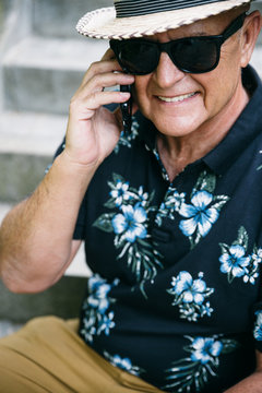 Stylish senior man smiling, wearing a straw hat and sunglasses, talking on the phone and sitting on stairs