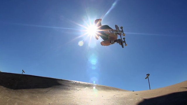 A Skateboarder jumps out of a Skatepark bowl through sun burst and lands.