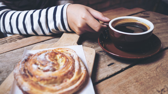 Closeup image of a raisin danish and a woman holding and drinking hot coffee on wooden vintage table in coffee shop