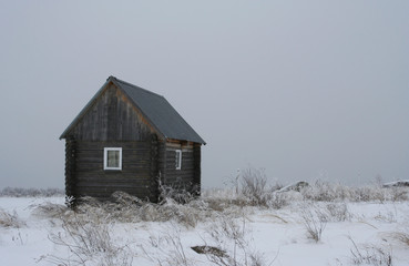 The small wooden house on the snow-covered field with dried grass on the cloudy winter day. 