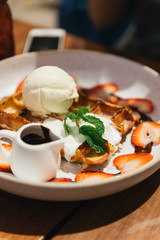Waffle topping with a scoop of vanilla ice cream, whip cream, sliced strawberry, mint leaves and chocolate sauce on white plate at dessert cafe in Bangkok, Thailand.