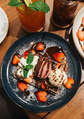 Chocolate waffle topping with scoop of vanilla and strawberry ice cream, whip cream, sliced strawberry, mint leaves, icing and chocolate sauce on blue plate at dessert cafe in Bangkok, Thailand.