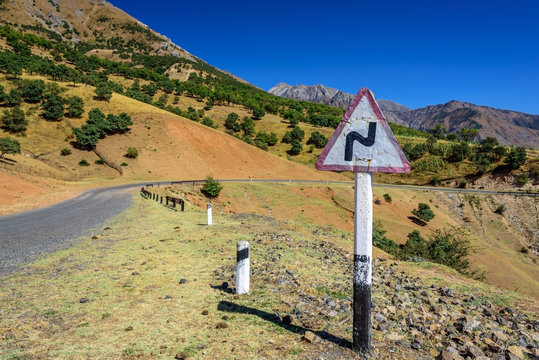 Curved road warning sign on the side of the serpentine road on the background of the mountains covered with trees and blue sky
