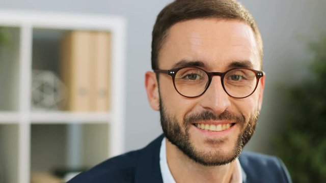 Close-up portrait of successful young businessman in glasses looking straight to the camera and smiling, feeling happy and satisfied. Indoor shot.