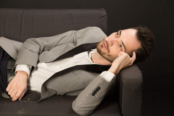 Fototapeta premium portrait of a young man with an unshaven face in a suit with a pensive face lying on a sofa against a dark background