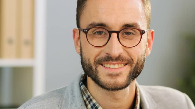Close up face of an attractive caucasian man in spectacles, wearing a casual shirt and a grey shirt sitting at the office desk, looking and smiling at the camera.Indoor shot.