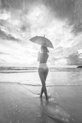 Back view of beautiful topless woman with umbrella standing in the rain on the beautiful beach over cloudy sky background - black and white photo