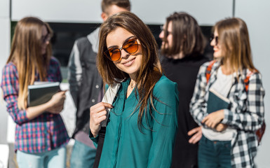 Sunny day portrait of cheerful brunette girl with backpack wears green shirt in sunglasses before group of students