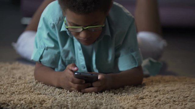 African american kid playing in application on phone comfortably lying on floor