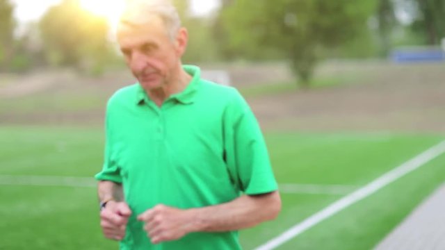 Man note down the time in his watch and start running. Senior man runs near football field. Active elderly man in green t-shirt runs around a football field.Physical activity helps to burn up calories