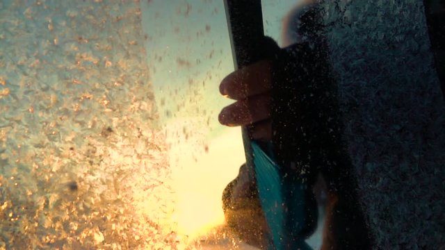 SLOW MOTION CLOSE UP DOF: Person cleaning morning frost off a car window at golden sunrise. Hand scraping hoarfrost off a frozen car windshield, snowflakes flowing over the sun. Scraper removing frost