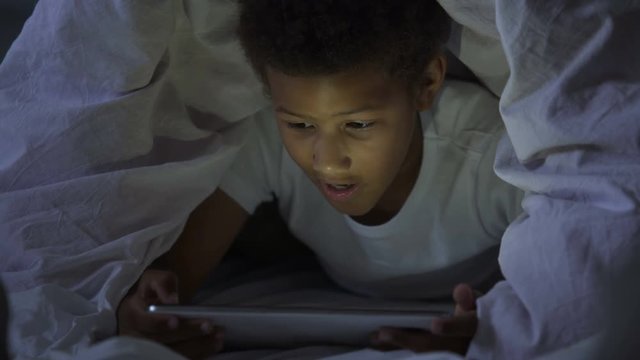 Little kid looking at tablet secretly at night, covered with head in blanket