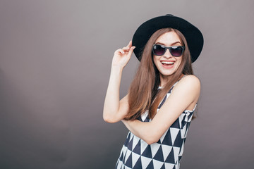 Young beautiful fashionable woman with trendy makeup in black hat and glasses on the grey background . Model looking at camera, wearing stylish eyeglasses. Female fashion, beauty concept.

