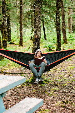Young Man Sitting In A Hammock Reading A Book In the Woods