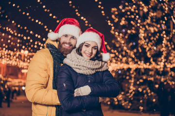Close up focused portrait of cute family cuddling in the town while on stroll at x mas evening, wearing santa`s head wear, warm outfits, gloves, many sparkling lights on the background
