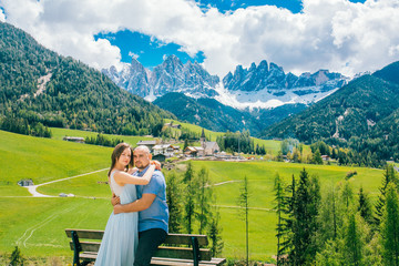 Fototapeta na wymiar Young couple admires the view in relaxation. Famous best alpine place. Santa Maddalena village with magical Dolomites mountains in background, Val di Funes valley, Trentino Alto Adige Italy