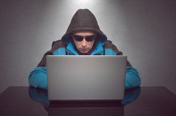 Hacker. Young man in a jacket with a hood and sun glasses working on laptop computer on the table isolated. Network security.