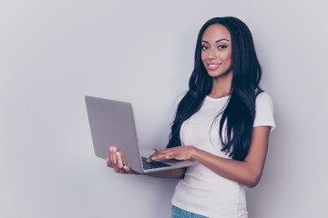 Naklejka premium Portrait of successful afro lady with laptop, typing on a keyboard, standing on pure gray backgrund, looking in the camera, She is in a casual smart with natural daily cosmetics, long hair