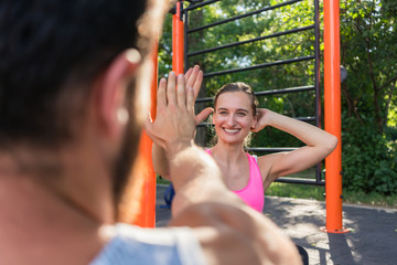 Obraz premium Over the shoulder view of a cheerful young woman giving high-five to her partner while doing crunches outdoors, during couple workout in a fitness park