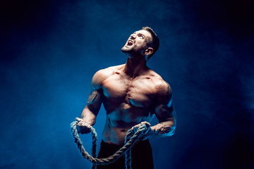 Side view of muscular bodybuilder man training hard with rope in studio, smoke.