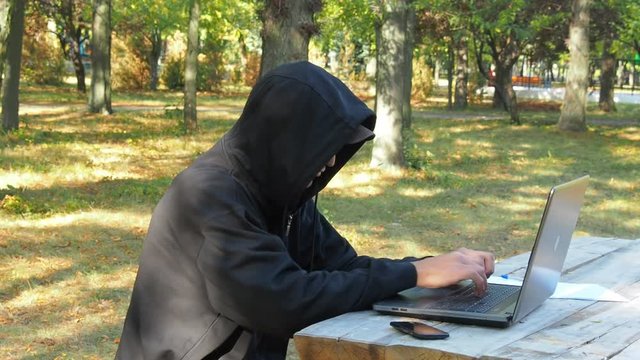 A hacker works with a laptop in the park. A man in a hood with a laptop outdoors.