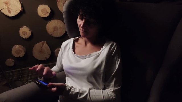 Relaxed woman using smartphone sitting in armchair under the lamp 