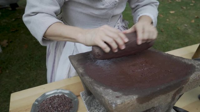 Woman in 18th century dress using a stone grinder, mane and metate to grind cocoa beans into chocolate.