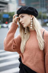 A beautiful young blonde woman dressed in a large warm pink sweater with a black beret and gorgeous earrings. She goes through a big spectacular Chinese city in autumn.