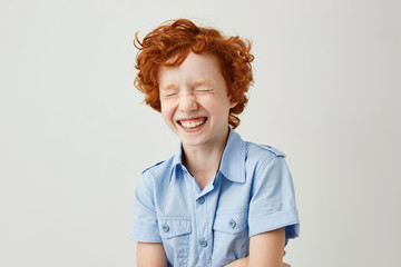 Close up portrait of cheerful little kid with curly ginger hair and freckles laughing with closed...