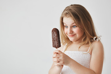 Close up of beautiful light-haired small girl with blue eyes in white dress holding ice-cream in hands, looking at it with happy face expression.