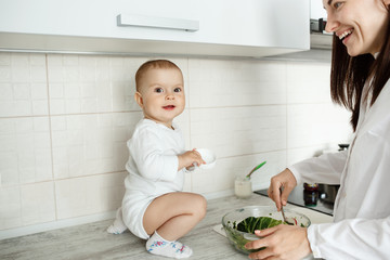 Portrait of cheerful little baby boy sitting on kitchen table, smiling and trying to help mother with cooking salad while mom brightfully smiling.