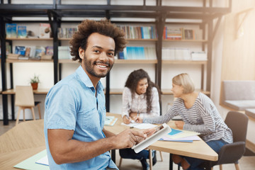 Close up portrait of good-looking black-skinned university student sitting on meeting with friends after study, talking about group project, looking through websites on digital tablet.