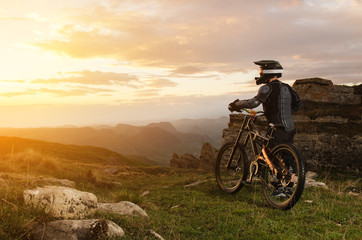 The rider in full protective equipment on the mtb bike is riding toward the sunset in the rays of the sunset sun against the background of the rocks of the setting sun and clouds