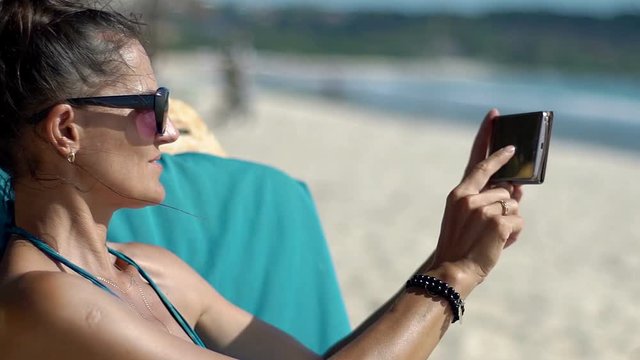 Beautiful woman sitting on the sandy beach and doing photos on smartphone, steadycam shot
