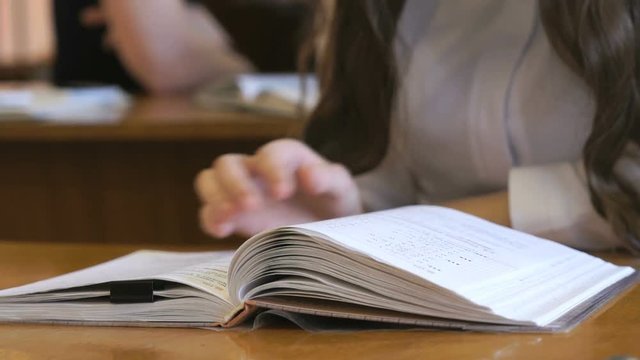 Schoolgirl dressed in white shirt sitting at school desk flips the pages of the school book. Close up