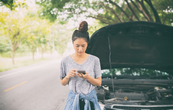 Asian Woman Using Mobile Phone While Looking And Stressed Man Sitting After A Car Breakdown On Street