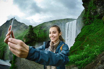 Naklejka premium smiling woman in warm clothes making selfie on a Skogafoss waterfall background, Iceland. Focus on the woman