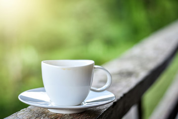 White cup of coffee on wooden railing by the green garden.