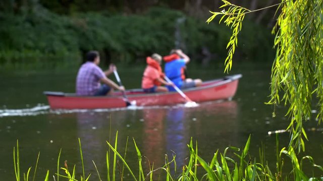 Focus on foreground tree and grasses as adult with boy and girl paddle past in a red kayak on a large pond surrounded by trees. Side view in summer sunlight, 4K