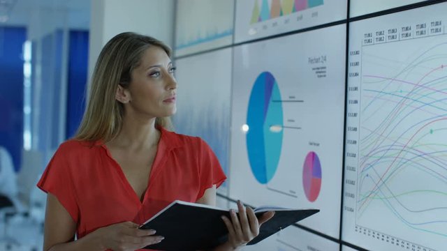  Portrait smiling businesswoman standing next to video wall with graphs & data