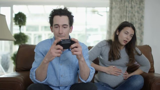  Competitive man playing video games at home, with bored pregnant wife